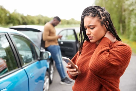 Young woman stands outside of a car holding her neck in pain and using her cell phone after an accident