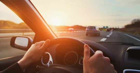 Close up of a young person's hands on the steering wheel while driving on the highway at sunset